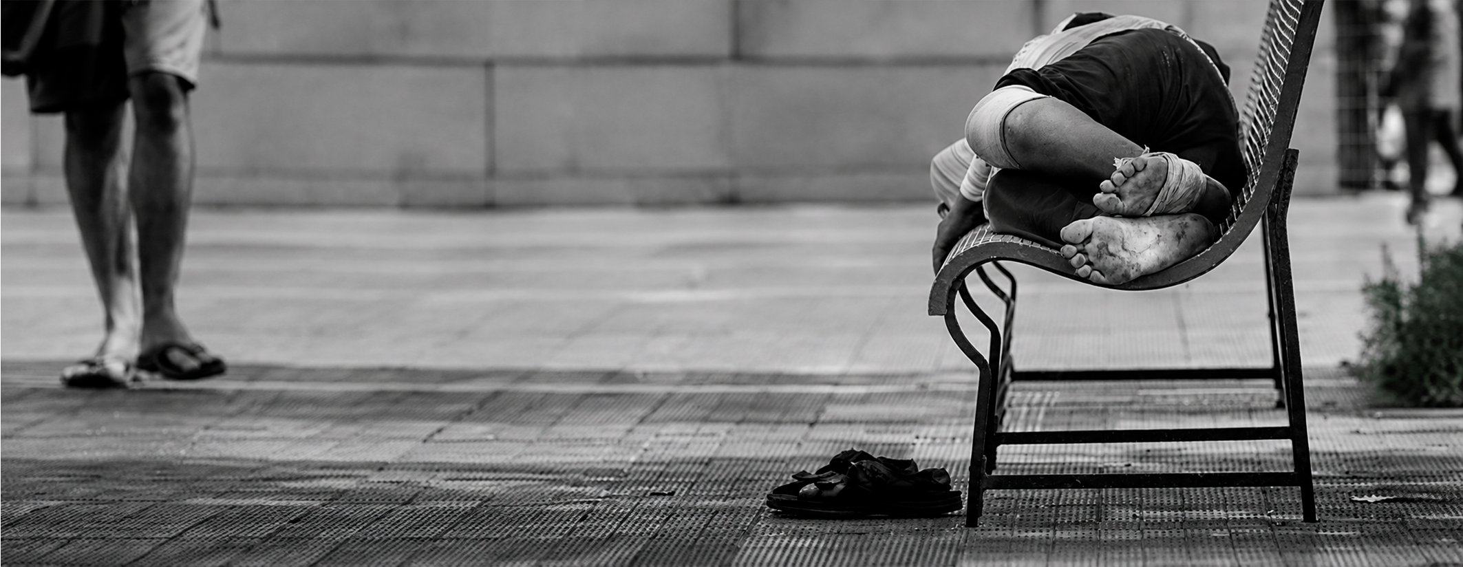 Una persona en situación de calle duerme sobre un banco público, con sus zapatillas apoyadas en el piso junto al banco.
