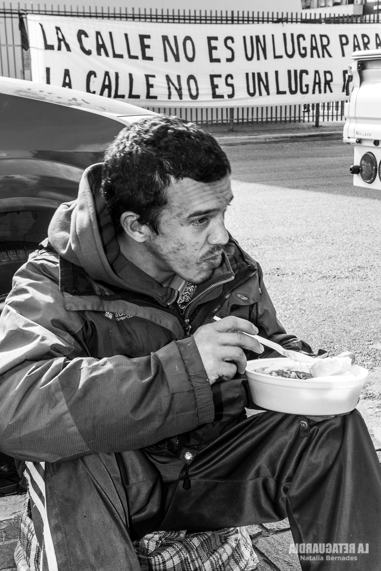 Un hombre en situación de calle come de un recipiente, sentado en la vía pública frente a un cartel que dice "La calle no es un lugar para vivir". Foto: Natalia Bernades / La Retaguardia.